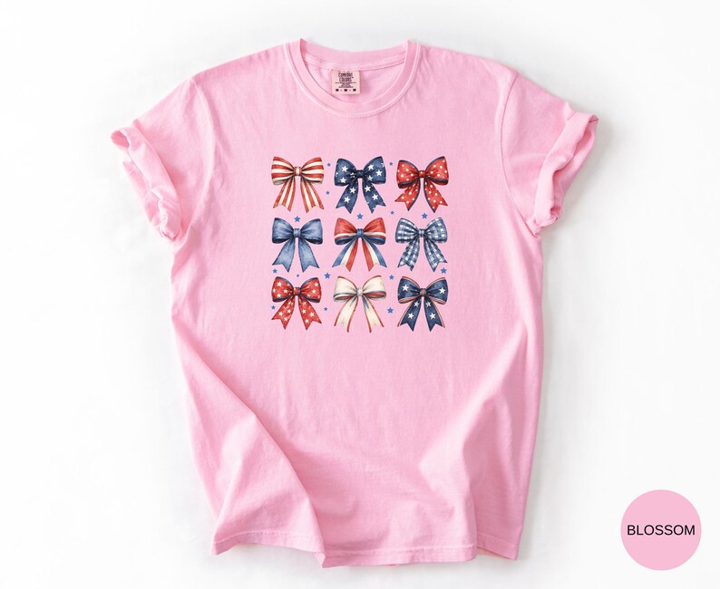 May include: A light pink short-sleeved t-shirt featuring a central graphic print of nine decorative bows arranged in a grid. The bows display various red, white, and blue patterns including stripes, polka dots, and stars, accented by small blue stars.