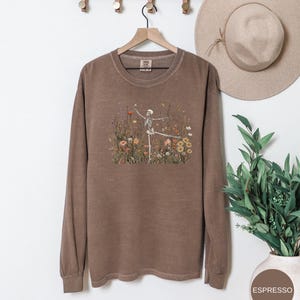 Puede incluir: Una sudadera de manga larga con cuello redondo en color marr&oacute;n apagado. El centro del pecho presenta un estampado gr&aacute;fico de un esqueleto bailando en medio de un campo de flores silvestres coloridas, incluyendo flores naranjas, amarillas y blancas con follaje verde.