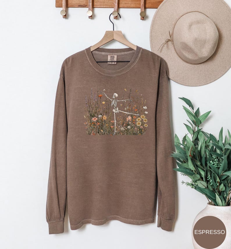 Puede incluir: Una sudadera de manga larga con cuello redondo en color marr&oacute;n apagado. El centro del pecho presenta un estampado gr&aacute;fico de un esqueleto bailando en medio de un campo de flores silvestres coloridas, incluyendo flores naranjas, amarillas y blancas con follaje verde.