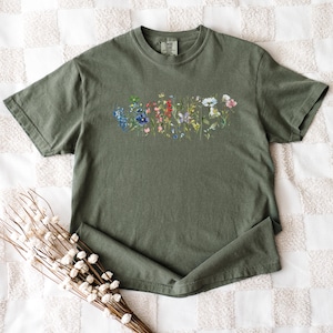 a green t-shirt with a floral design printed on it, placed on a white and beige checkered surface. Next to the t-shirt is a bunch of white flowers.