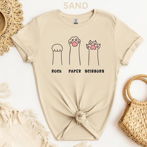 Puede incluir: Camiseta color arena con un dise&ntilde;o divertido de patas de gato que representan piedra, papel y tijeras. El texto "ROCK PAPER SCISSORS" est&aacute; impreso debajo de las ilustraciones de las patas. La camiseta est&aacute; hecha de material suave y tiene un cuello redondo cl&aacute;sico.