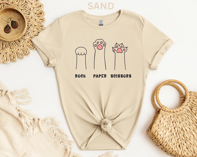 Puede incluir: Camiseta color arena con un dise&ntilde;o divertido de patas de gato que representan piedra, papel y tijeras. El texto "ROCK PAPER SCISSORS" est&aacute; impreso debajo de las ilustraciones de las patas. La camiseta est&aacute; hecha de material suave y tiene un cuello redondo cl&aacute;sico.