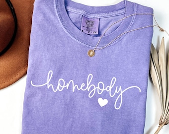 Puede incluir: Una camiseta violeta Comfort Colors con la palabra "homebody" en escritura blanca y un pequeño corazón blanco. También se ven un collar dorado y un sombrero marrón.