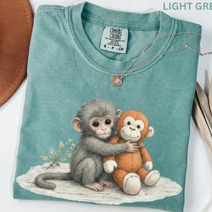 Puede incluir: Camiseta verde claro con un gr&aacute;fico de un mono gris abrazando a un mono de peluche naranja. La camiseta tiene cuello redondo y mangas cortas. Un collar plateado con un peque&ntilde;o colgante est&aacute; sobre la camiseta.