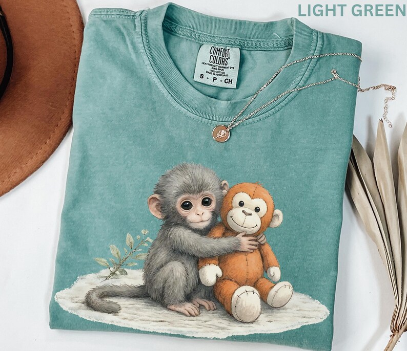 Puede incluir: Camiseta verde claro con un gr&aacute;fico de un mono gris abrazando a un mono de peluche naranja. La camiseta tiene cuello redondo y mangas cortas. Un collar plateado con un peque&ntilde;o colgante est&aacute; sobre la camiseta.