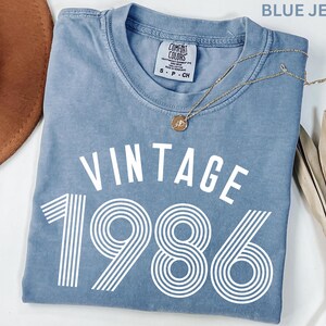 May include: A blue jean-colored t-shirt with the word "VINTAGE" above the number "1986" in white. A gold necklace with a pendant rests on the shirt. The shirt is a Comfort Colors brand.