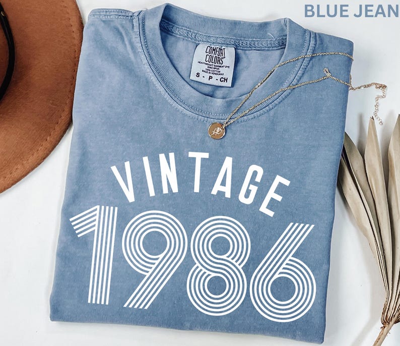 May include: A blue jean-colored t-shirt with the word "VINTAGE" above the number "1986" in white. A gold necklace with a pendant rests on the shirt. The shirt is a Comfort Colors brand.