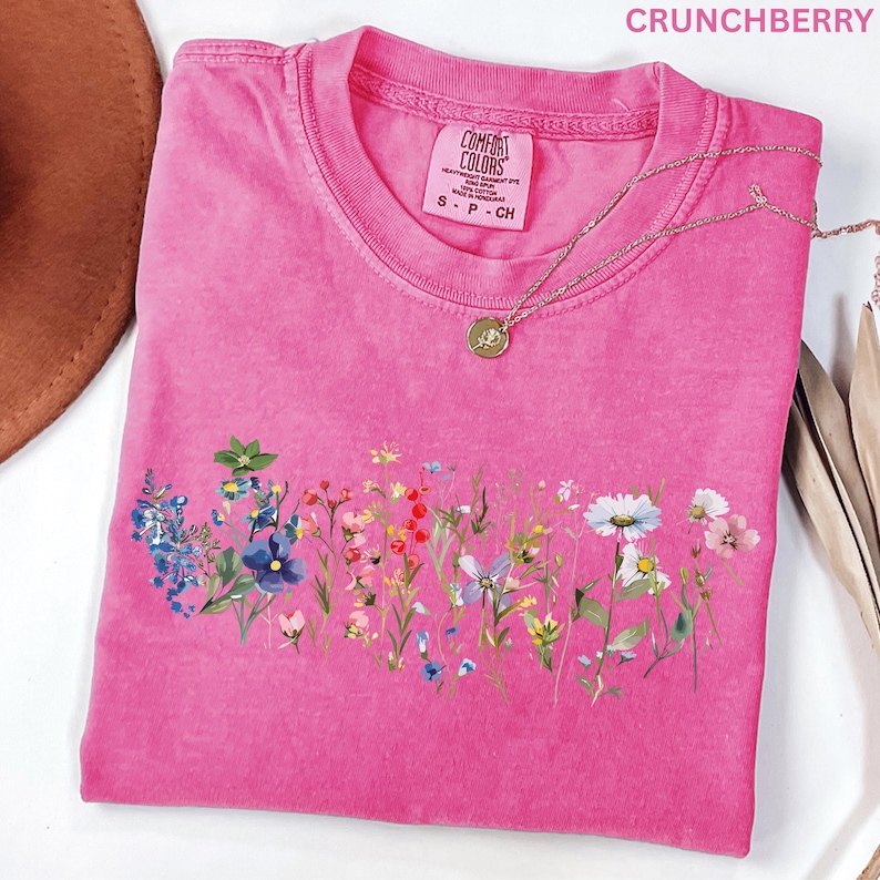 May include: A pink t-shirt with a floral design across the front. The shirt has a crew neck and short sleeves. A gold necklace with a round pendant is draped across the shirt. The word "CRUNCHBERRY" is printed at the top.