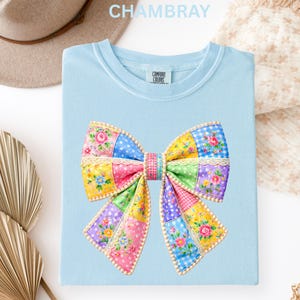 May include: Pale blue t-shirt featuring a vibrant patchwork bow design. The bow incorporates floral, polka dot, and gingham patterns in various colours. The shirt is made of a soft material and has the text "CHAMBRAY" at the top.