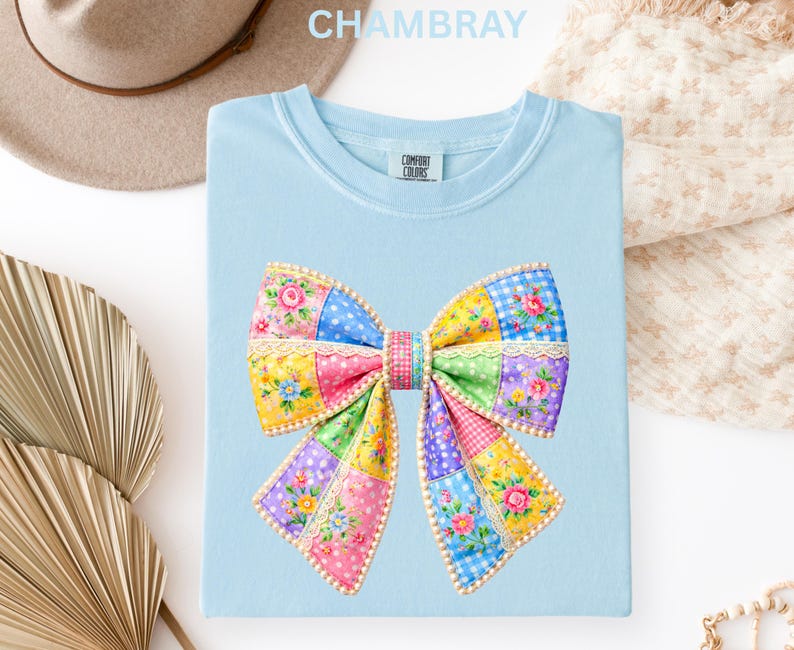 May include: Pale blue t-shirt featuring a vibrant patchwork bow design. The bow incorporates floral, polka dot, and gingham patterns in various colours. The shirt is made of a soft material and has the text "CHAMBRAY" at the top.