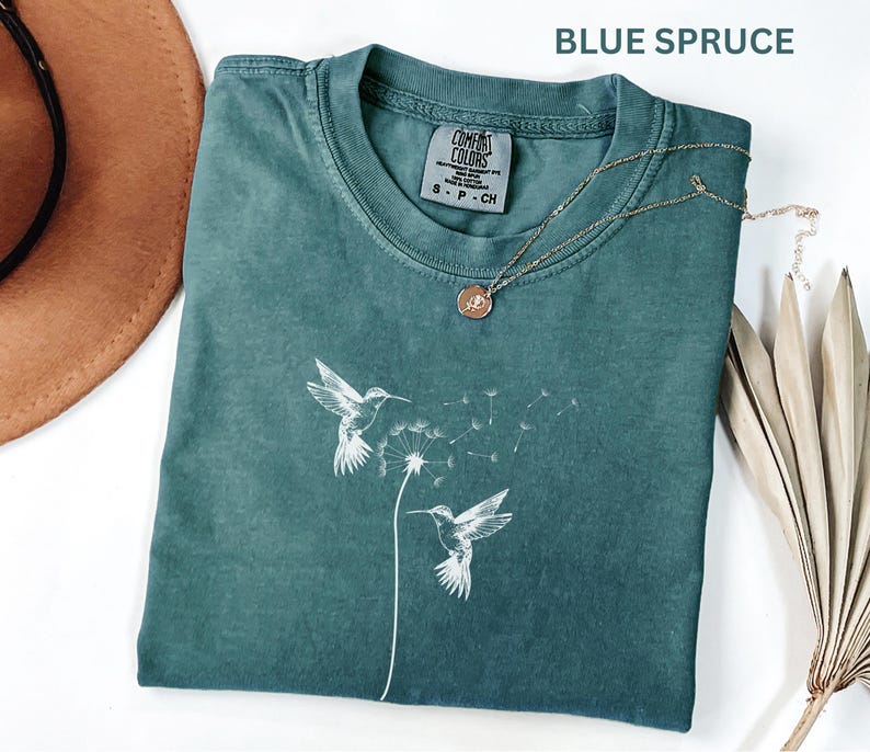 a green t-shirt with a white bird design, a brown hat, and a necklace with a pendant, all placed on a white surface.