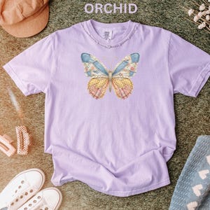 May include: A light purple t-shirt with a distressed neckline. The shirt features a butterfly design with blue, yellow, and pink patchwork wings. The word "ORCHID" is printed at the top of the image.