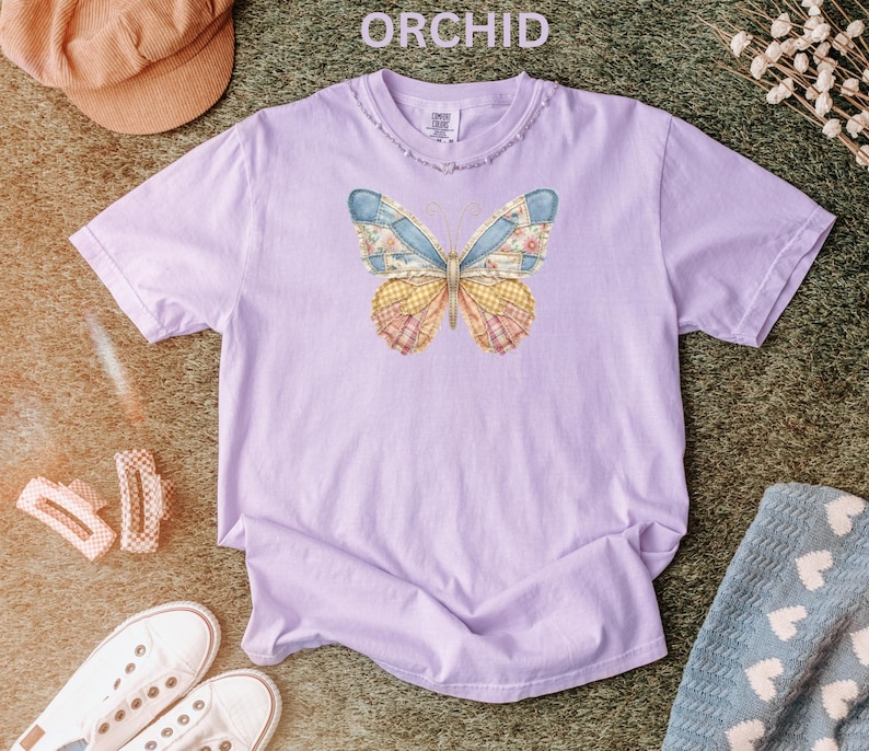 May include: A light purple t-shirt with a distressed neckline. The shirt features a butterfly design with blue, yellow, and pink patchwork wings. The word "ORCHID" is printed at the top of the image.
