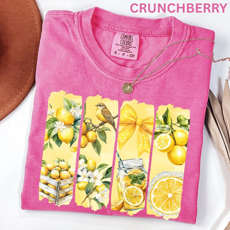 May include: A pink t-shirt with a lemon-themed graphic. The graphic features yellow lemons, a bird, a bow, and a mason jar with lemons. The shirt has a crew neck and short sleeves. The word "CRUNCHBERRY" is printed at the top.