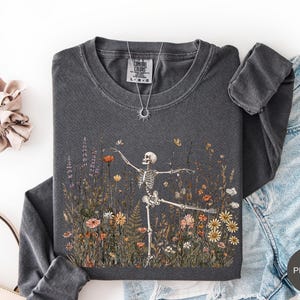 Puede incluir: Una camiseta de manga larga gris carb&oacute;n con un gr&aacute;fico de un esqueleto bailando en un campo de flores silvestres coloridas. La camiseta tiene cuello redondo y una etiqueta Comfort Colors en el interior. Se incluye un collar con dije de sol.