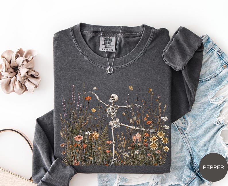 Puede incluir: Una camiseta de manga larga gris carb&oacute;n con un gr&aacute;fico de un esqueleto bailando en un campo de flores silvestres coloridas. La camiseta tiene cuello redondo y una etiqueta Comfort Colors en el interior. Se incluye un collar con dije de sol.