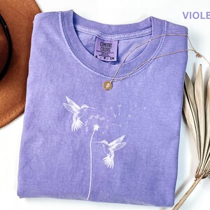 a purple t-shirt with a white design of two hummingbirds, a necklace, a hat, and a fan on a white background.