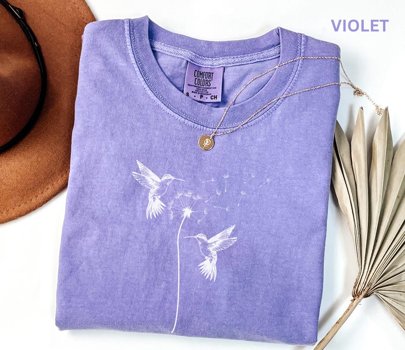 a purple t-shirt with a white design of two hummingbirds, a necklace, a hat, and a fan on a white background.