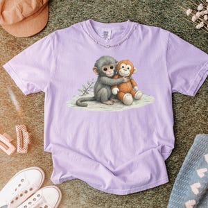 Puede incluir: Una camiseta de color lila claro con una ilustraci&oacute;n de acuarela de un mono gris abrazando a un mono de peluche naranja. El dise&ntilde;o est&aacute; en una camiseta lila claro con un cuello desgastado.