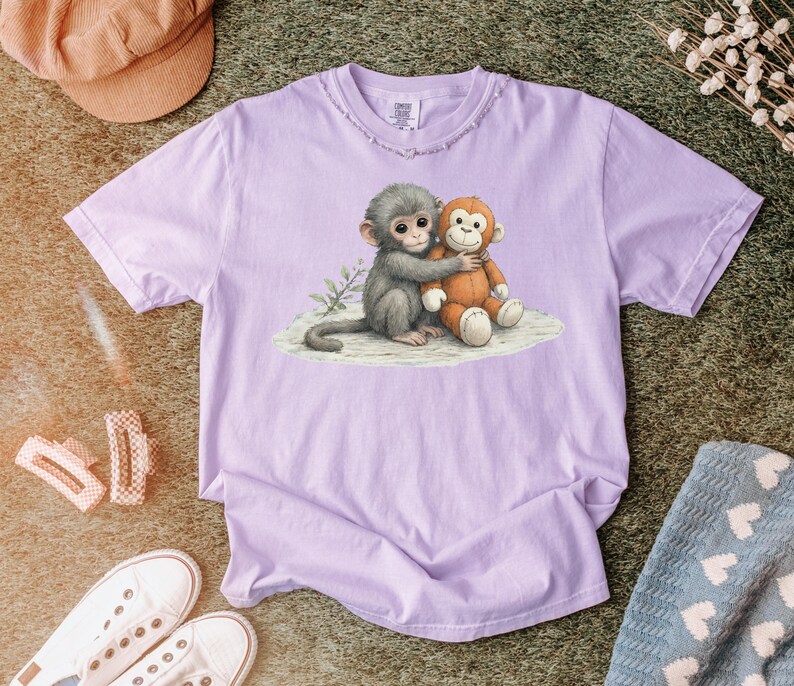Puede incluir: Una camiseta de color lila claro con una ilustraci&oacute;n de acuarela de un mono gris abrazando a un mono de peluche naranja. El dise&ntilde;o est&aacute; en una camiseta lila claro con un cuello desgastado.