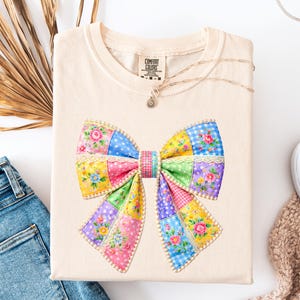 May include: A cream-coloured t-shirt with a large, colourful bow design. The bow is made of patchwork squares in various colours, including pink, yellow, blue, and green, with floral and polka dot patterns. The shirt is neatly folded.