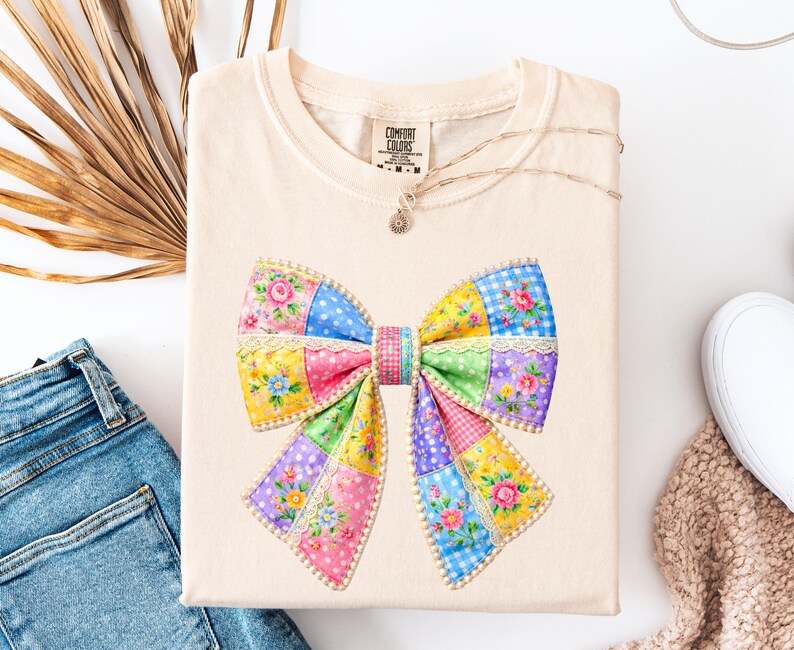 May include: A cream-coloured t-shirt with a large, colourful bow design. The bow is made of patchwork squares in various colours, including pink, yellow, blue, and green, with floral and polka dot patterns. The shirt is neatly folded.