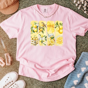 May include: Pink t-shirt with a lemon-themed graphic. The graphic features yellow vertical panels with lemon illustrations, including lemons in a crate, a bird on a branch, a yellow bow, and a jar of lemonade. The shirt is surrounded by accessories.