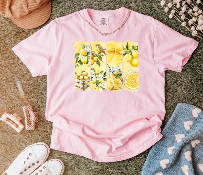 May include: Pink t-shirt with a lemon-themed graphic. The graphic features yellow vertical panels with lemon illustrations, including lemons in a crate, a bird on a branch, a yellow bow, and a jar of lemonade. The shirt is surrounded by accessories.