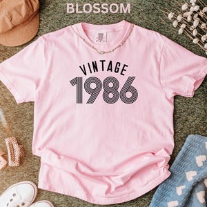 May include: A light pink t-shirt with the text "VINTAGE 1986" in black. The shirt is short-sleeved and has a round neckline. The word "BLOSSOM" is at the top of the image. A necklace is around the neckline.