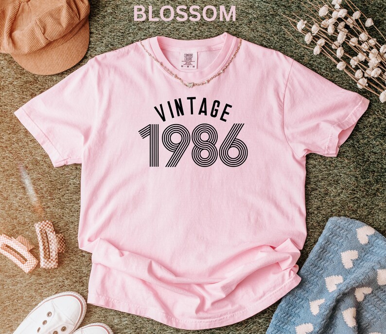 May include: A light pink t-shirt with the text "VINTAGE 1986" in black. The shirt is short-sleeved and has a round neckline. The word "BLOSSOM" is at the top of the image. A necklace is around the neckline.