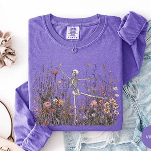 Puede incluir: Una camiseta violeta de manga larga con un estampado gr&aacute;fico de un esqueleto bailando en un campo de flores silvestres de colores. La camiseta tiene cuello redondo y una etiqueta de la marca Comfort Colors visible en el cuello. Tela texturizada.