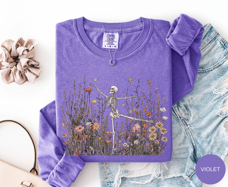 Puede incluir: Una camiseta violeta de manga larga con un estampado gr&aacute;fico de un esqueleto bailando en un campo de flores silvestres de colores. La camiseta tiene cuello redondo y una etiqueta de la marca Comfort Colors visible en el cuello. Tela texturizada.