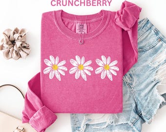 May include: A long-sleeve crewneck t-shirt in a vibrant pink color featuring three white daisy graphics with yellow centers across the chest. The shirt has a Comfort Colors label at the inner neckline and is displayed with denim shorts and a scrunchie.