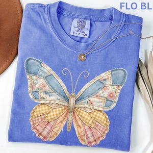 May include: A blue t-shirt with a patchwork butterfly design. The butterfly wings feature denim, floral, and plaid patterns. A gold necklace with a pendant is draped across the shirt. The text "FLO BLUE" is at the top.