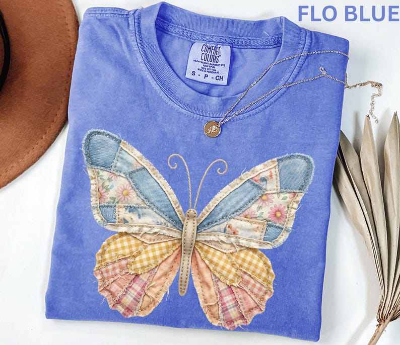 May include: A blue t-shirt with a patchwork butterfly design. The butterfly wings feature denim, floral, and plaid patterns. A gold necklace with a pendant is draped across the shirt. The text "FLO BLUE" is at the top.