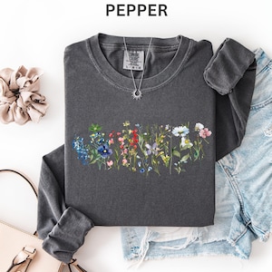 May include: A dark gray long-sleeve shirt with a floral design in shades of blue, red, pink, and white. The shirt is paired with denim shorts, a light pink purse, and a beige scrunchie. A silver necklace with a sun pendant is also visible.