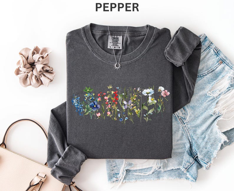 May include: A dark gray long-sleeve shirt with a floral design in shades of blue, red, pink, and white. The shirt is paired with denim shorts, a light pink purse, and a beige scrunchie. A silver necklace with a sun pendant is also visible.