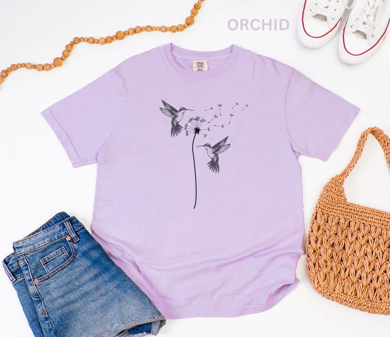 a casual outfit consisting of a purple t-shirt with a graphic design, blue jeans, a woven straw bag, and a pair of sneakers.