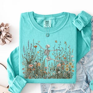Puede incluir: Una sudadera de manga larga color verde menta con cuello redondo que presenta un estampado gr&aacute;fico de un esqueleto bailando en medio de un campo de flores silvestres coloridas y mariposas. La etiqueta interior indica Comfort Colors, talla L.