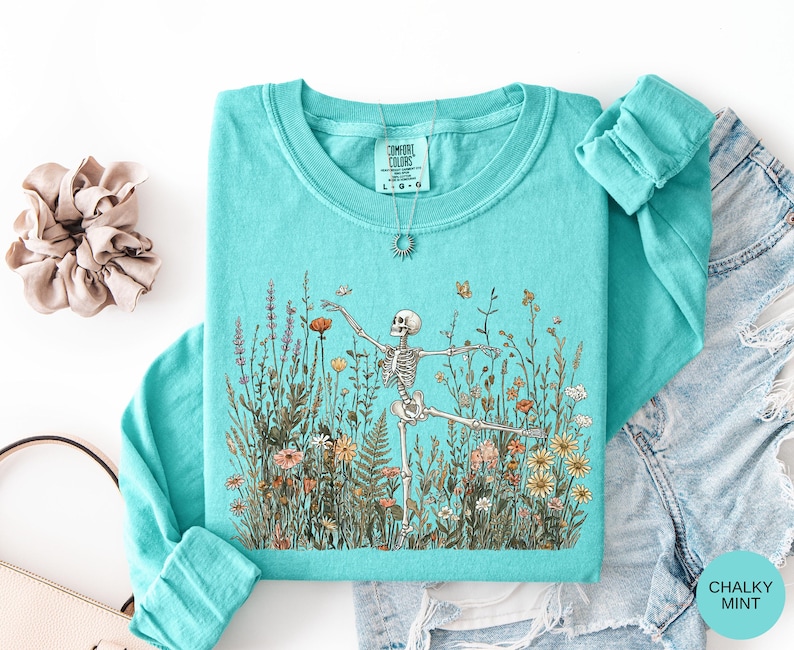Puede incluir: Una sudadera de manga larga color verde menta con cuello redondo que presenta un estampado gr&aacute;fico de un esqueleto bailando en medio de un campo de flores silvestres coloridas y mariposas. La etiqueta interior indica Comfort Colors, talla L.