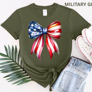 May include: Military green t-shirt featuring a watercolor bow design in red, white, and blue. The bow has stars and stripes, evoking patriotic themes. The shirt is paired with denim shorts and white sneakers, suggesting casual wear.