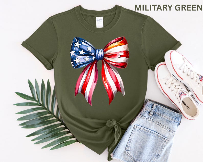 May include: Military green t-shirt featuring a watercolor bow design in red, white, and blue. The bow has stars and stripes, evoking patriotic themes. The shirt is paired with denim shorts and white sneakers, suggesting casual wear.