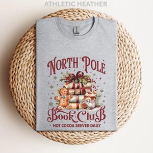 A gray t-shirt with a Christmas-themed design, including a gingerbread man, presents, and a hot chocolate mug, along with the text "North Pole Book Club" and "Hot Cocoa Served Daily".