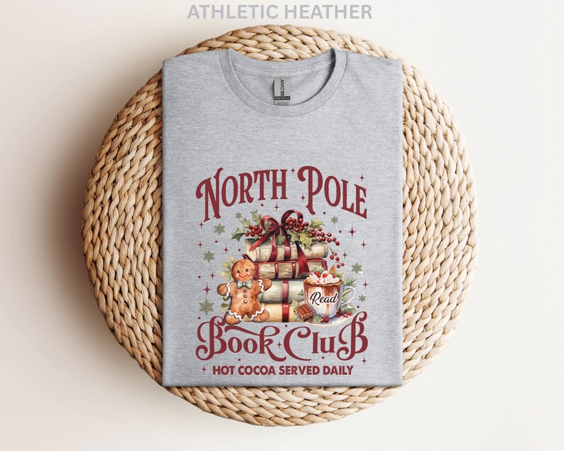 A gray t-shirt with a Christmas-themed design, including a gingerbread man, presents, and a hot chocolate mug, along with the text "North Pole Book Club" and "Hot Cocoa Served Daily".