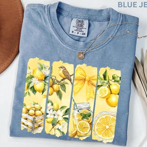 May include: Blue jean Comfort Colors t-shirt with a lemon-themed graphic. The graphic features lemons, a bird, a yellow bow, and a jar of lemonade. A gold necklace is draped across the shirt.