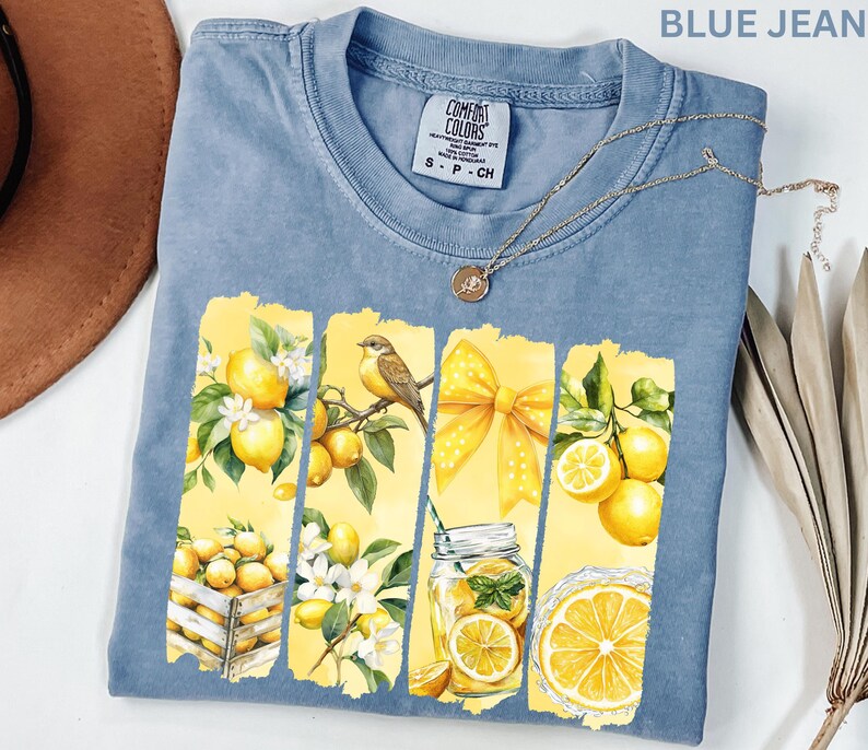 May include: Blue jean Comfort Colors t-shirt with a lemon-themed graphic. The graphic features lemons, a bird, a yellow bow, and a jar of lemonade. A gold necklace is draped across the shirt.