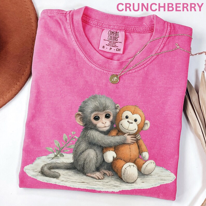 Puede incluir: Una camiseta rosa Comfort Colors con un gr&aacute;fico de dos monos abraz&aacute;ndose. Un mono es gris y el otro es un juguete de peluche naranja. La camiseta tiene cuello redondo y una etiqueta peque&ntilde;a. La palabra "CRUNCHBERRY" est&aacute; impresa en la parte superior.