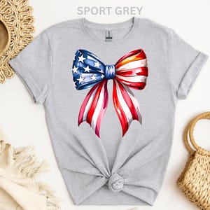 May include: Sport grey t-shirt featuring a watercolor bow design in red, white, and blue, with stars and stripes. The bow is centered on the front of the shirt. The text "SPORT GREY" is at the top.