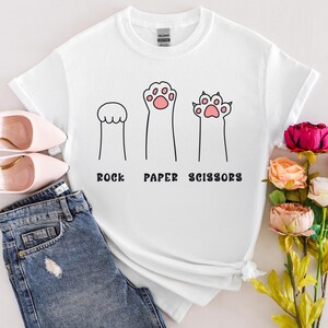 Puede incluir: Camiseta blanca con un dise&ntilde;o l&uacute;dico de "Piedra, Papel o Tijeras" con ilustraciones de patas de gato. El dise&ntilde;o incluye tres figuras en forma de pata, con el texto debajo. La camiseta se combina con vaqueros azules y zapatos rosas.