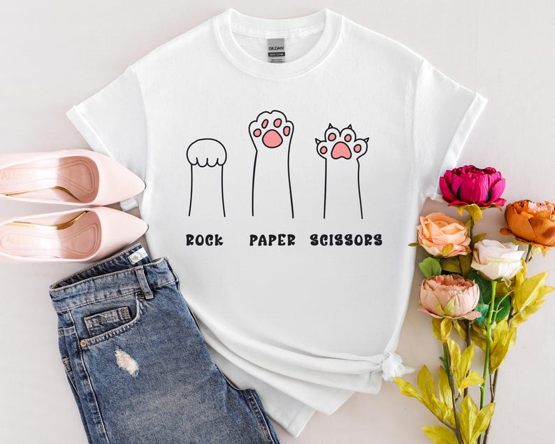 Puede incluir: Camiseta blanca con un dise&ntilde;o l&uacute;dico de "Piedra, Papel o Tijeras" con ilustraciones de patas de gato. El dise&ntilde;o incluye tres figuras en forma de pata, con el texto debajo. La camiseta se combina con vaqueros azules y zapatos rosas.